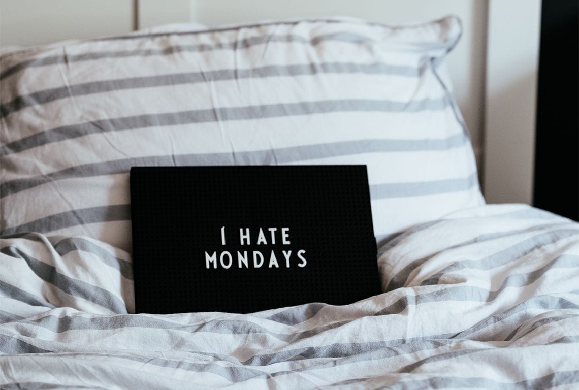 A black pillow on a bed that says I hate Mondays in white letters